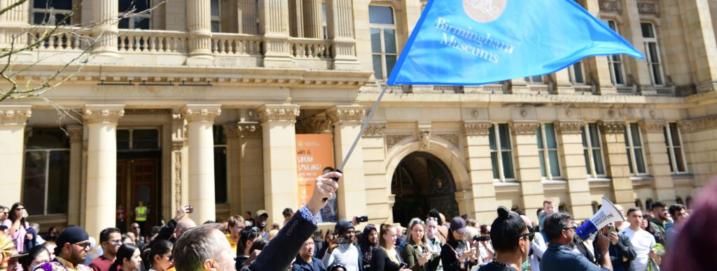 Outside shot of the a museum with a crowd of people in front of it. One person is holding up a blue flag with the Birmingham Museums Trust logo.